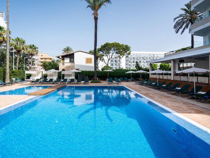 Piscina al aire libre clara con tumbonas en un hotel con palmeras y cielo despejado.