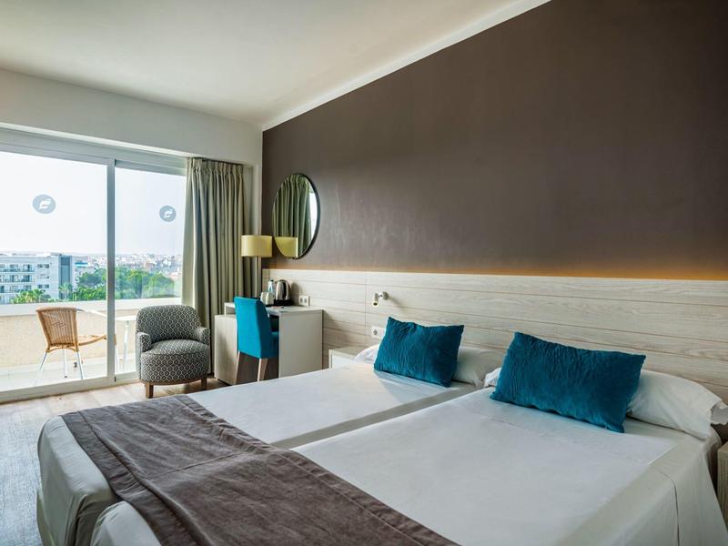 Habitación de hotel moderna con dos camas, cojines azules y balcón con sillas.