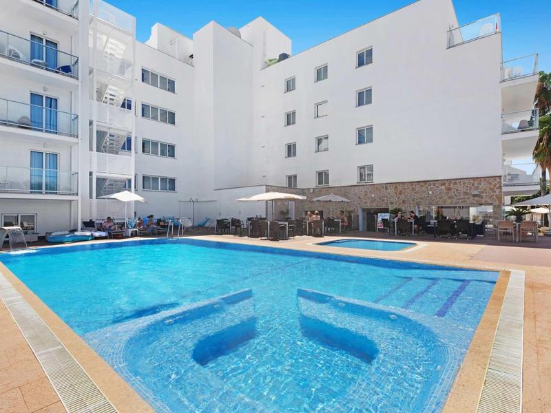 Piscina clara con asientos y sombrillas junto a un edificio de hotel blanco bajo un cielo azul.