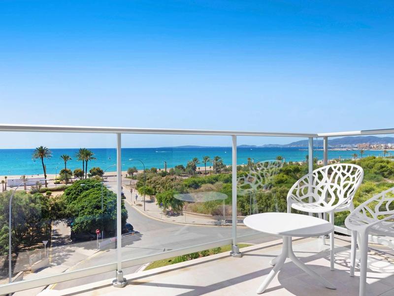 Balcón con sillas y mesa blancas con vista al mar y palmeras bajo cielo azul.