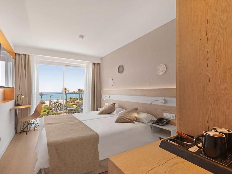 Habitación de hotel moderna con cama doble, balcón y vista al mar con luz diurna.
