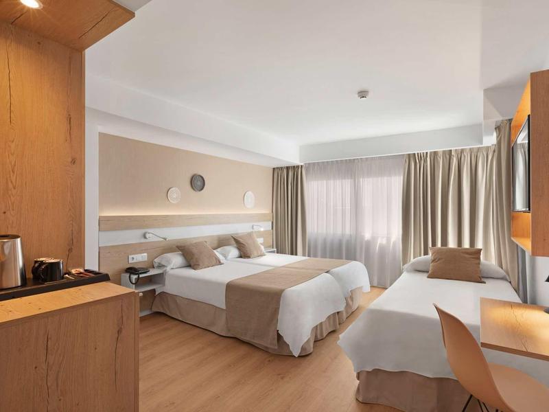 Habitación de hotel luminosa con tres camas, paneles de madera y una gran ventana con cortinas.