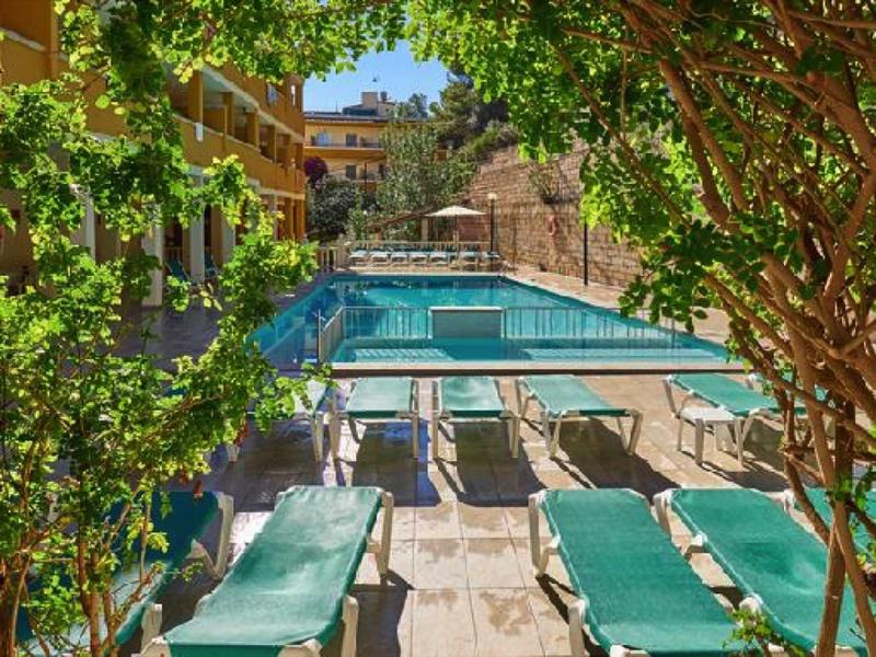 Vue d'une piscine avec des chaises longues vertes et des arbres, entourée de bâtiments.