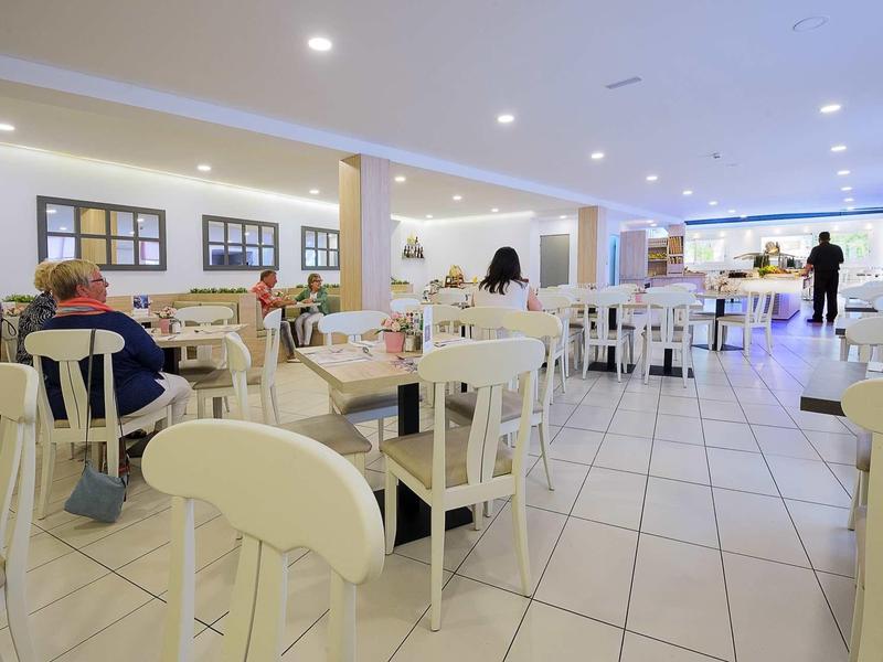 Salle à manger lumineuse avec des chaises et tables blanches, plusieurs invités et des lumières de plafond visibles.