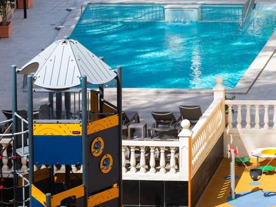 Strefa basenowa z placem zabaw dla dzieci i leżakami, otoczona murami.