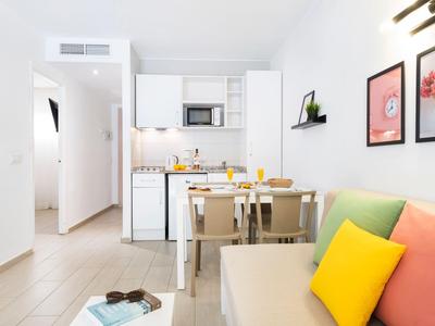 Jasny salon z aneksem kuchennym, stołem jadalnym i kolorowymi poduszkami na sofie.