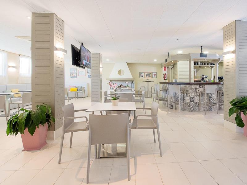 Jasna, nowoczesna sala śniadaniowa z białymi stołami i krzesłami oraz barem w tle.