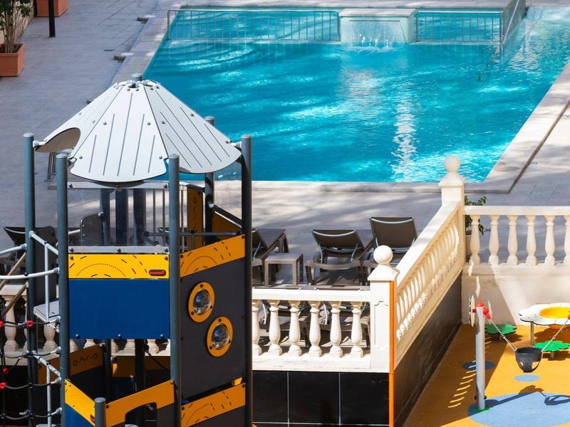 Strefa basenowa z placem zabaw dla dzieci i leżakami, otoczona murami.