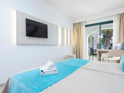 Jasny pokój hotelowy z niebieskimi akcentami, łóżkiem podwójnym, telewizorem i balkonem z widokiem na palmy.