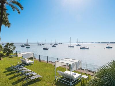 Blick auf ruhige Bucht mit Segelbooten, grüner Wiese, Liegen und zwei weißen Sonnendecks bei Sonnenschein.