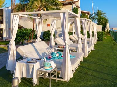Weiße Strandbetten mit Vorhängen auf grünem Rasen neben einem Hotelgebäude unter blauem Himmel.