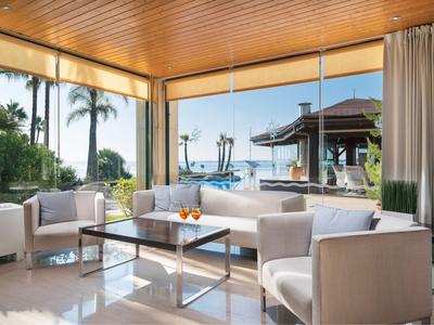 Helle Lounge mit großen Fenstern, weißen Sesseln und Blick auf ein tropisches Außenbecken.