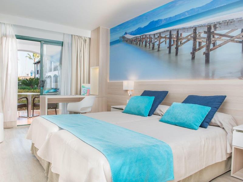 Nowoczesny pokój hotelowy z łóżkiem podwójnym, niebieskimi akcentami i widokiem na morze z balkonu.