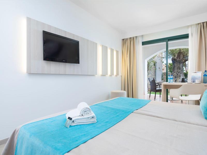 Jasny pokój hotelowy z niebieskimi akcentami, łóżkiem podwójnym, telewizorem i balkonem z widokiem na palmy.