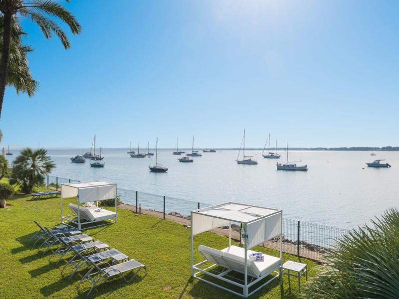 Widok na słoneczny ogród z leżakami i białymi łóżkami przy wodzie z żaglówkami.