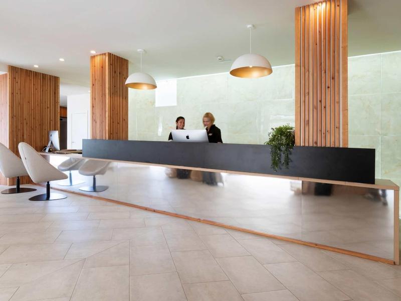 Modern hotel reception with long black counter and wooden columns, two receptionists.