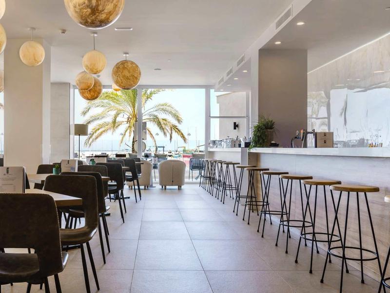 Bright modern hotel bar with high stools, dining tables, and palm view through large windows.