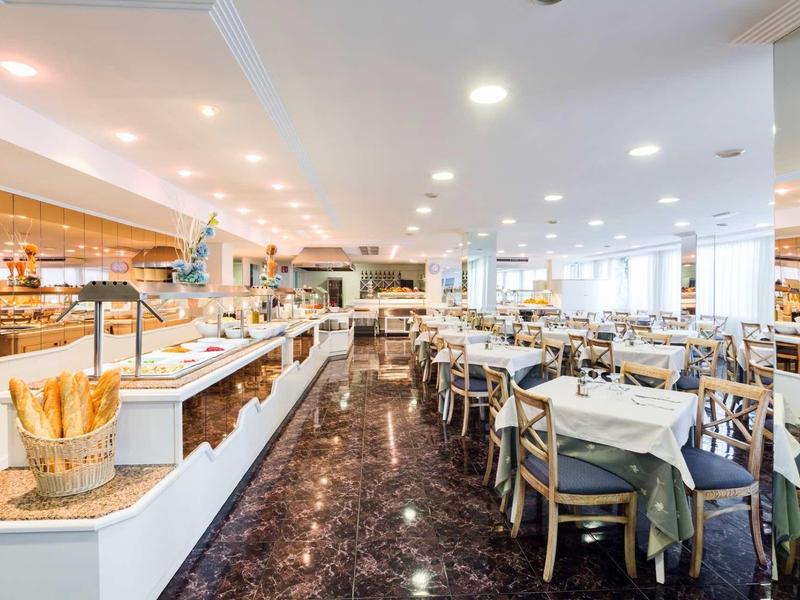 Helles Restaurant mit vielen Tischen, gedeckten Stühlen und Buffetbereich links, helle Decke.