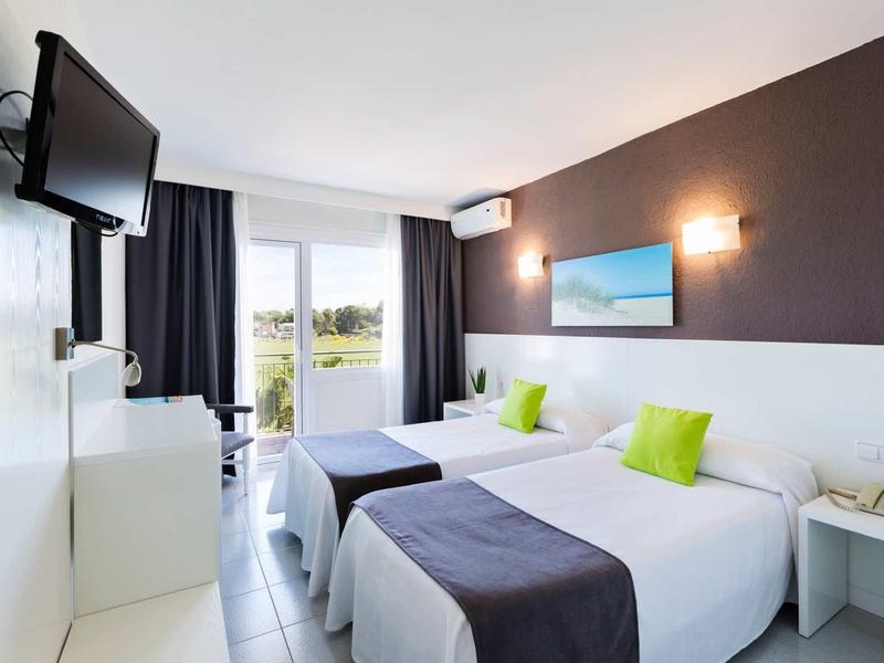 Helle Doppelzimmer mit zwei Betten, grünen Kissen, Fernseher und Balkon mit Vorhängen.