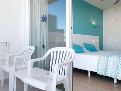 Hotelzimmer mit Bett, bunten Decken und zwei weißen Plastikstühlen auf dem Balkon.