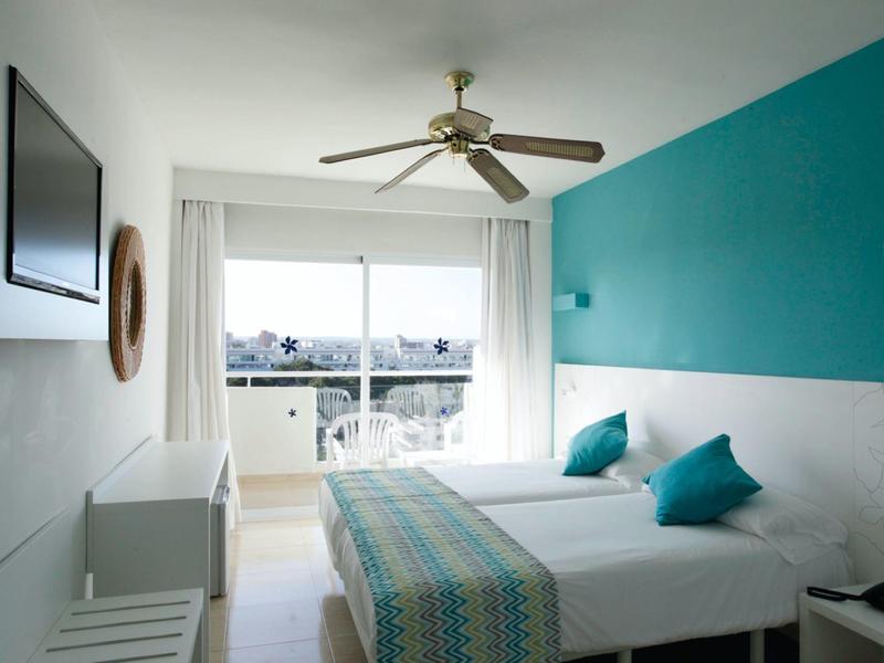 Camera d'hotel luminosa con due letti singoli, cuscini turchesi e vista mare dal balcone.