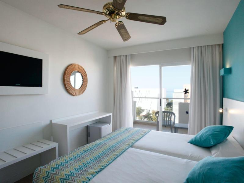 Camera d'hotel luminosa con due letti, ventilatore a soffitto, TV e balcone con vista sul mare.