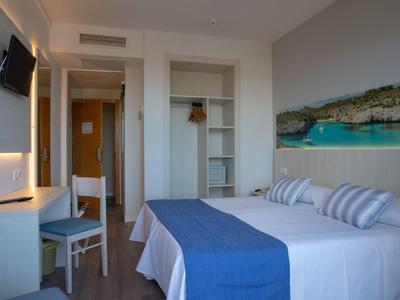 Helles Hotelzimmer mit Doppelbett, blauer Bettdecke, Schreibtisch, Stuhl und Meerblickbild an der Wand.