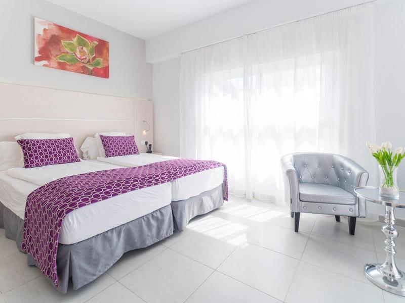 Chambre d'hôtel lumineuse avec lit double, accents violets sur le lit et coin salon près de la fenêtre.