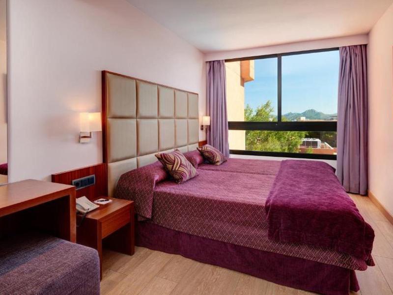 Modernes Hotelzimmer mit lila Bettdecke, großem Fenster und Blick auf Berge.