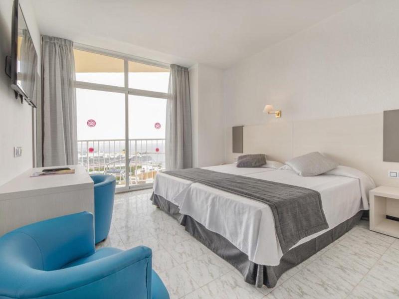 Moderní hotelový pokoj s manželskou postelí, modrým křeslem a balkonem s výhledem na moře.