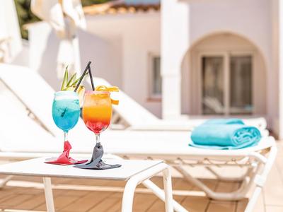 Zwei bunte Cocktails auf einem Tisch neben weißen Liegestühlen am sonnigen Poolbereich.