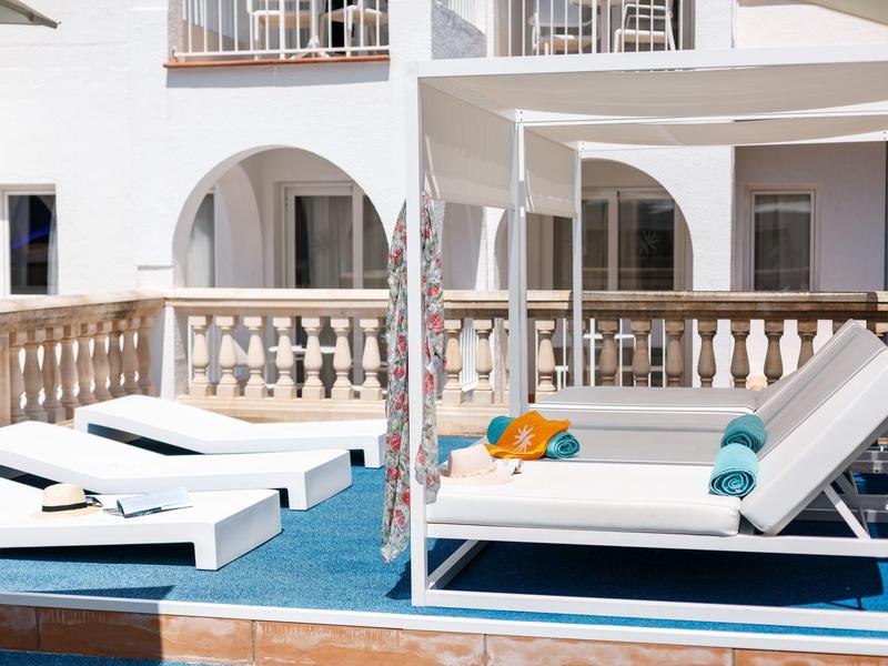 Witte ligstoelen en zonnescherm op een blauwe terras voor een hotel met witte muren en balkons.
