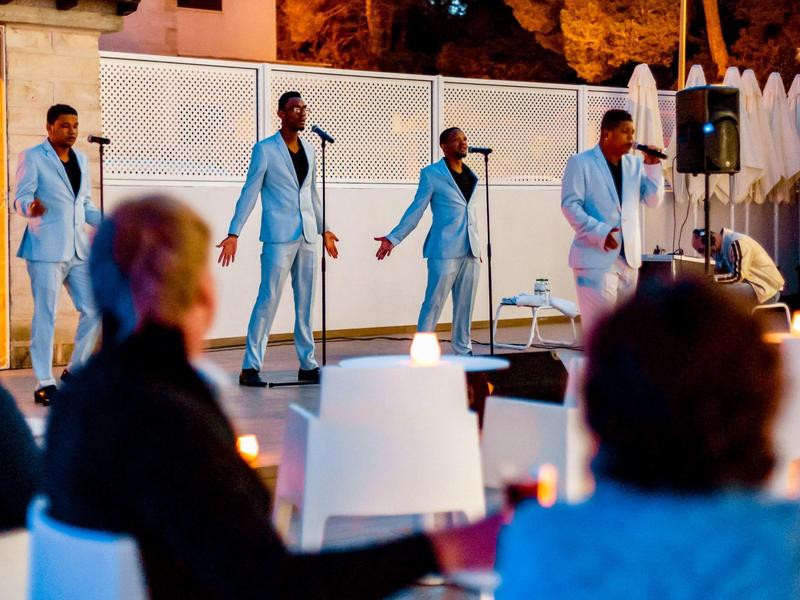 Vier mannen in blauwe pakken zingen op het podium voor zittend publiek.