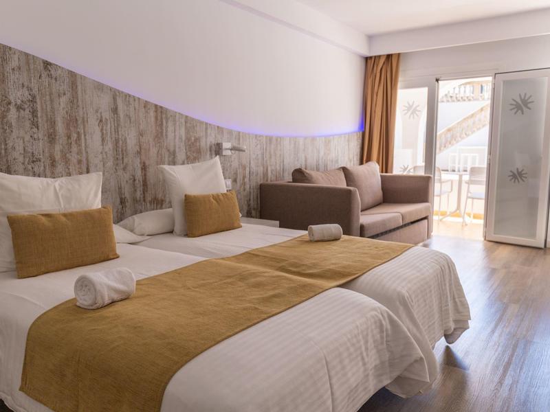 Modern hotelkamer met twee eenpersoonsbedden, bank en balkon met daglicht.