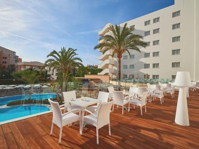 Terrasse mit weißen Stühlen und Tischen, Holzboden, Pool und Palmen vor mehrstöckigem Gebäude.