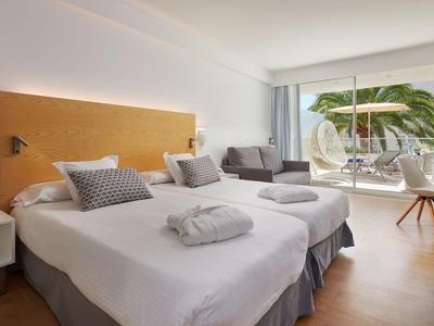 Modernes Hotelzimmer mit zwei Einzelbetten, hellen Holzmöbeln und Balkon mit Pflanzen und Sitzgelegenheit.