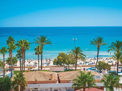 Strand mit Palmen, Strandliegen und blauem Meer unter klarem Himmel im Vordergrund Häuser mit roten Ziegeln.