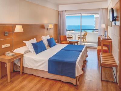 Helles Hotelzimmer mit Doppelbett, Holzmöbeln, blau-weißer Bettwäsche und Balkon mit Meerblick.