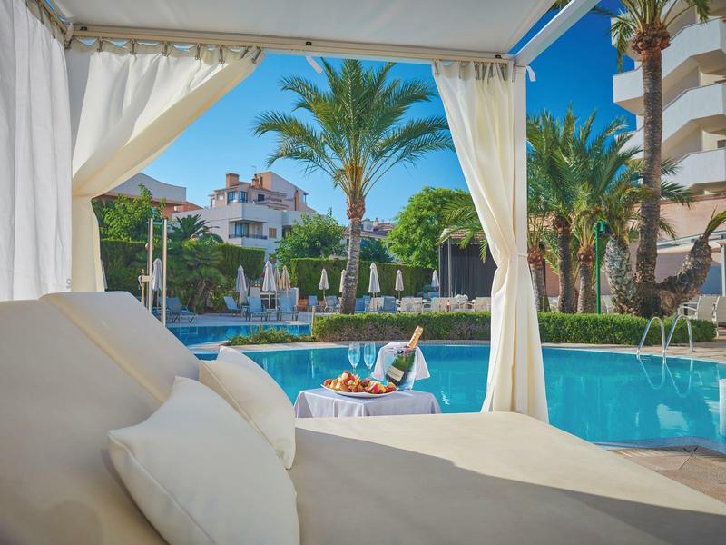 Gazebo rilassante a bordo piscina con tende, lettino e vista su palme e edifici dell'hotel.