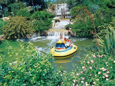 Zwei Personen fahren in einem gelben Boot auf einem Teich mit Wasserfall und blühenden Pflanzen.
