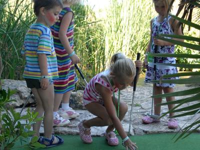 Vier Kinder spielen Minigolf auf einem grünen Spielfeld, umgeben von Pflanzen und Sonnenlicht.