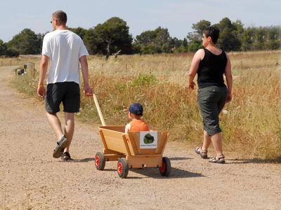 Zwei Erwachsene ziehen und schieben ein Kind im Holzwagen auf einem Feldweg.