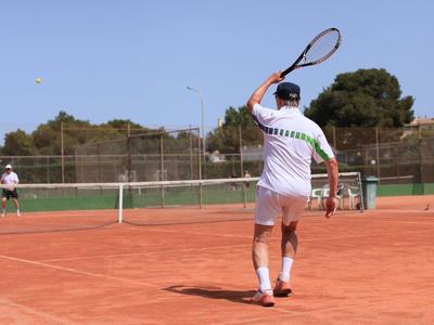 Zwei Tennisspieler auf rotem Sandplatz, einer bereitet einen Rückhandschlag vor, klarer Himmel.