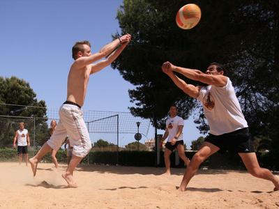 Zwei Männer spielen Beachvolleyball im Sand, umgeben von Bäumen und blauem Himmel.