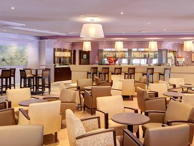 Großer, heller Hotel-Loungebereich mit beigen Sesseln, Holztischen und Bartresen im Hintergrund.