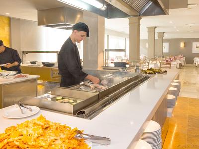 Moderne Restaurantküche mit Köchen, Grillplatte und Pizza auf hellem Tresen und Stühlen.