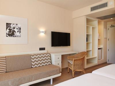 Helles Hotelzimmer mit Sofa, Wandbild, Fernseher, Schreibtisch, Stuhl und offener Kleiderschrank.