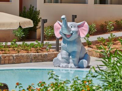 Bunter Elefantenbrunnen aus Kunststoff neben Pool, umgeben von Pflanzen und Gebäuden.