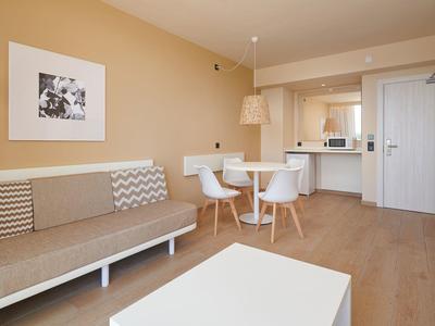 Moderner Wohnraum mit beiger Couch, rundem weißen Tisch, offener Küche und hellem Holzboden.