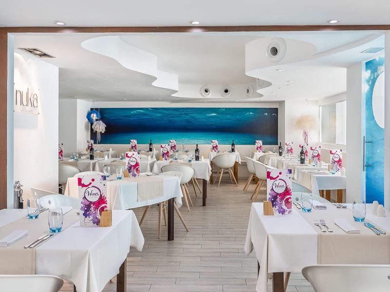 Elegantní restaurace s bílými stoly a velkým modrým mořským obrazem na stěně.